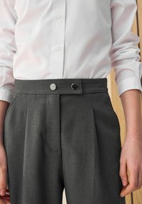 Dunkelgraue Hose mit Faltenwurf, die einen Knopfverschluss und kontrastierende schwarze und silberne Knopfdetails aufweist. Glatte Stofftextur.