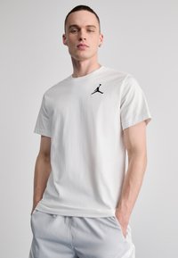 Fehér, rövid ujjú pamut póló; a mellkasán egy kis fekete Jumpman logó található. A modellt viselő személy a zsebébe dugja a kezét.