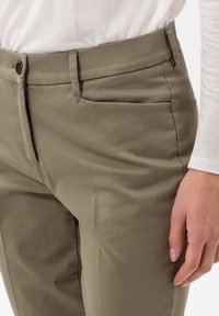 Zbliżenie na osobę ubraną w jasnobrązowe spodnie z guzikiem i zamkiem błyskawicznym oraz białą koszulę z długim rękawem, ręka oparta na boku.