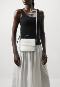Bolso cruzado de cuero blanco con un diseño estructurado, que presenta un asa superior y un logotipo en relieve. La modelo lleva un top negro ajustado y una falda blanca fluida.
