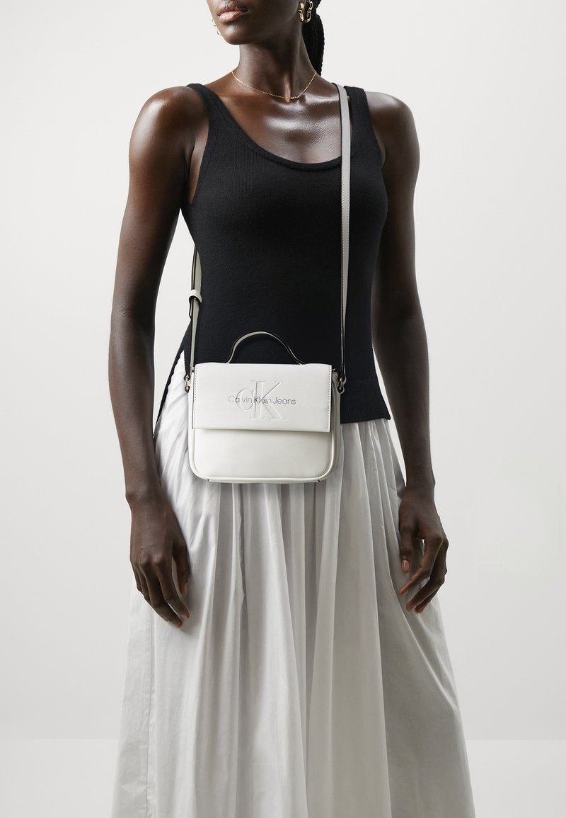 Bolso cruzado de cuero blanco con un diseño estructurado, que presenta un asa superior y un logotipo en relieve. La modelo lleva un top negro ajustado y una falda blanca fluida.