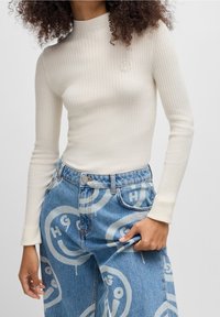 Ribbed, bluzka z długim rękawem w kolorze ecru, z subtelnym logo; w zestawieniu z niebieskimi jeansami z powtarzającym się wzorem graficznym.