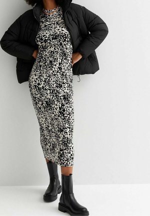 Osoba ubrana w czarną kurtkę puchową na czarno-białą sukienkę midi w kropki, do której dobrano czarne skórzane botki do połowy łydki, na białym tle.