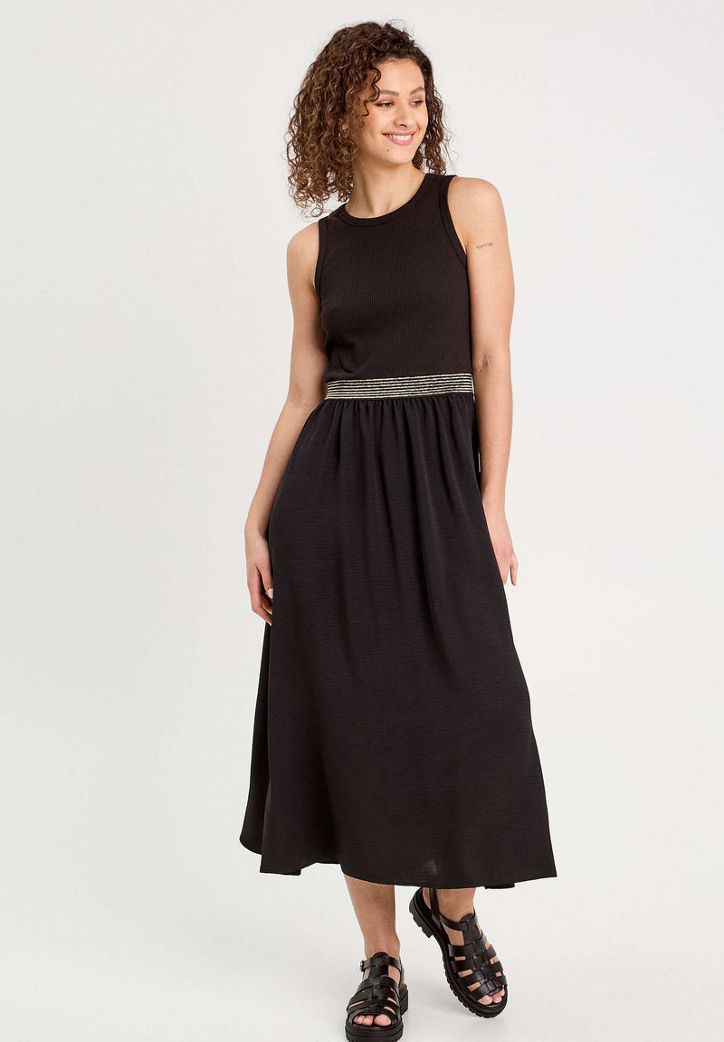 Robe maxi sans manches noire avec texture côtelée, jupe froncée et une ceinture décorative à rayures dorées. Associée à des sandales noires à lanières.