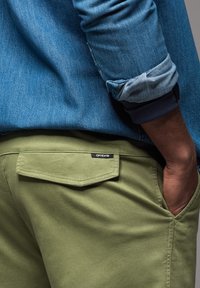 Olivgrüne Hose mit einer Gesäßtasche, auf der das Label "Ombre" zu sehen ist. Kombiniert mit einem Jeanshemd aus hellblauem Stoff und hochgekrempelten Ärmeln.