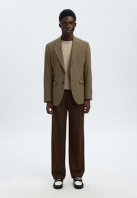 Άνδρας με καφέ καρό μπλέιζερ, μπεζ πουλόβερ, σκούρο καφέ παντελόνι με φαρδιά πόδια και μαύρα και λευκά μοκασίνια, στέκεται μπροστά σε μονόχρωμο φόντο.