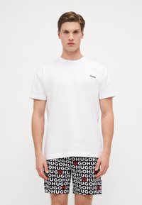 T-shirt en coton blanc avec le logo noir "HUGO" sur la poitrine, associé à un short noir présentant un motif de texte "HUGO" blanc et rouge répété.