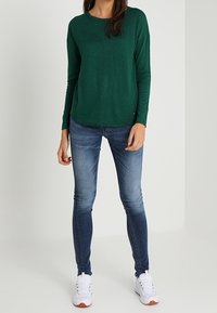 Grünes, langärmliges Oberteil aus leichtem Stoff, kombiniert mit figurbetonten blauen Jeans und weißen Sportschuhen mit orangefarbenen Akzenten.