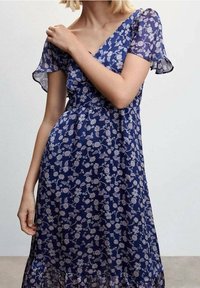 Robe florale marine avec des manches courtes et volantes. Présente un décolleté en V et des fronces à la taille. Fabriquée en tissu léger et transparent.