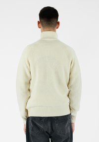 Maglione lavorato a maglia color crema con colletto alto, polsini e orlo a costine. Presenta un motivo testurizzato e dettagli cuciti sul retro.