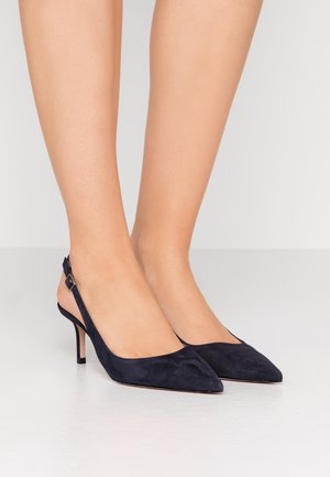 Ben iført sorte ruskind spidse slingback hæle med mellemhøj stilet på en glat hvid gulv mod en ensartet lys baggrund.