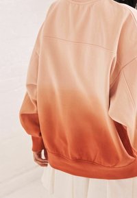 Oversized sweter z gradientem od jasnego brzoskwiniowego do głębokiego pomarańczowego, z prążkowanymi mankietami i dołem, wykonany z miękkiego materiału.