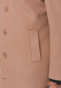 Abrigo de lana marrón claro con una textura lisa, con dos botones grandes y un bolsillo lateral. Diseño simple y ajuste entallado.