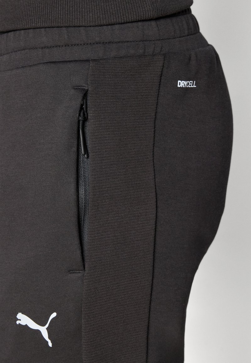 Pantalones deportivos negros con cintura de canalé y bolsillo con cremallera lateral. Presenta la etiqueta "DRYCELL" y el logotipo blanco de Puma en la pierna. Textura de tela suave.