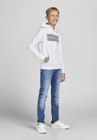 Felpa bianca con logo stampato in strisce nere e rosse, abbinata a jeans di denim azzurro chiaro e sneaker bianche. Abbigliamento semplice e casual.