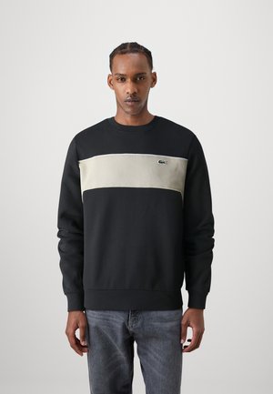 Sweat-shirt noir avec un panneau horizontal beige, des poignets côtelés et un petit logo vert sur la poitrine. Texture lisse, design à col rond.