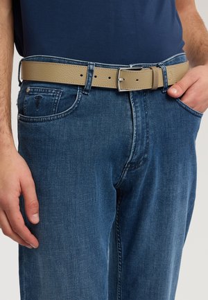 Man wearing blue denim jeans with a beige textured leather belt, left hand relaxed, right hand in pocket, navy blue shirt tucked in.