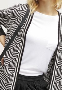 Černobílý vzorovaný kardigan s geometrickým designem, nošený přes obyčejné bílé tričko, doplněný černou plisovanou sukní.