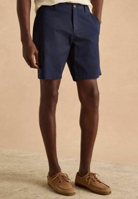 Shorts bleumarin, cu o croială clasică, realizate din bumbac. Dispune de buzunare laterale și închidere cu nasture. Asortați cu pantofi de bărbat din suede maromiu deschis.