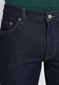 Tmavě modré džíny z denimu s měděnými detaily, praktickými kapsami a kontrastním šitím na švech. Detailní pohled na pas.