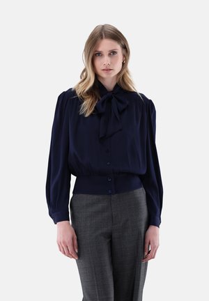 Frau trägt eine dunkelblaue Bluse mit Schleifenkragen und graue hoch taillierte Hose, stehend vor einem schlichten hellen Hintergrund.