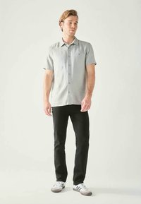 Camicia grigia con maniche corte e bottoni, in tessuto uniforme, design con collo, indossata con pantaloni neri e sneakers bianche, in piedi contro uno sfondo chiaro.
