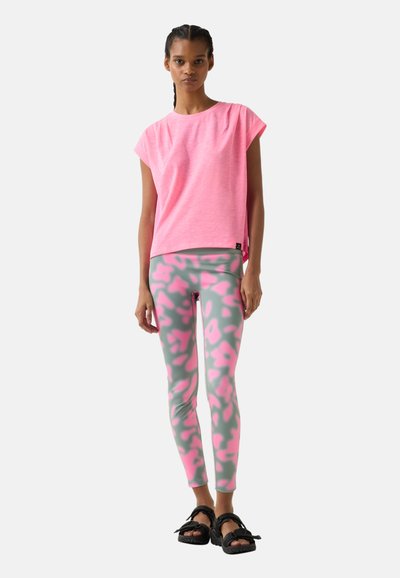 Junge Frau trägt ein pinkfarbenes Kurzarmoberteil, grau-pink gemusterte Leggings und schwarze Sandalen, steht vor einem einfarbigen Hintergrund.