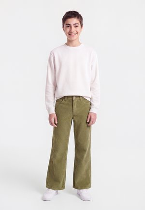 Glimlachende jongen staand, draagt een wit langarmshirt, olijfgroene brede ribcord broek en witte sneakers op een witte achtergrond.