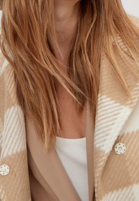 Dlouhé červené vlasy přes béžový kostkovaný kabát s velkými bílými knoflíky, béžový blazér a bílý top, viditelná pouze oblast krku a hrudníku.