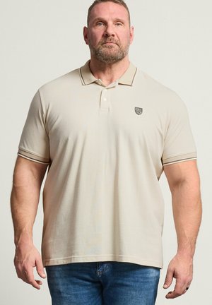 Mand med kort skæg iført beige polo skjorte med lille emblem og blå jeans, stående mod en ensartet lys baggrund.