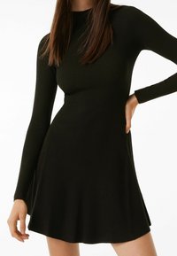 Robe noire côtelée à manches longues, corsage ajusté et jupe évasée. Comprend un col montant et des fentes latérales discrètes pour faciliter les mouvements.