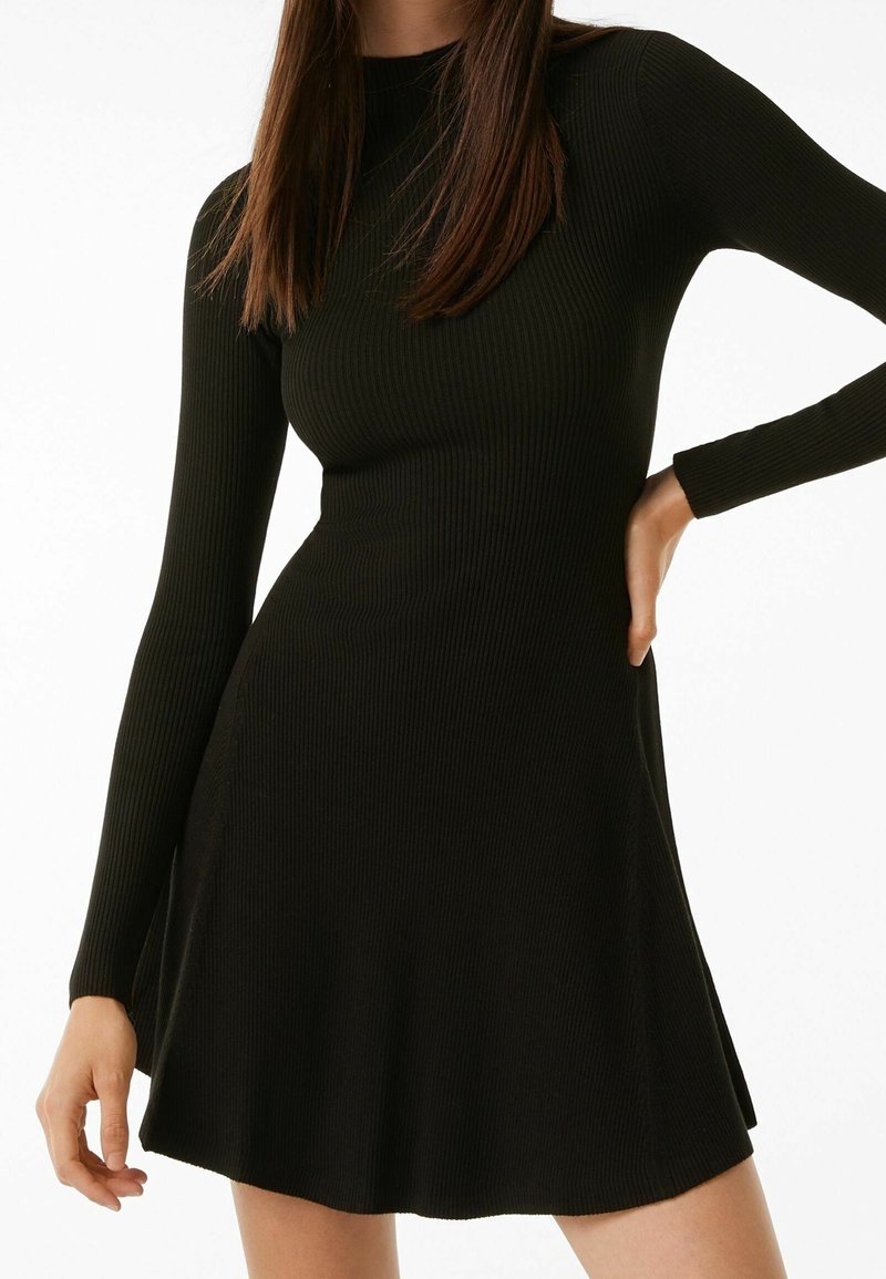 Robe noire côtelée à manches longues, corsage ajusté et jupe évasée. Comprend un col montant et des fentes latérales discrètes pour faciliter les mouvements.