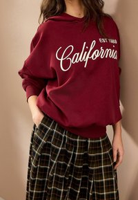 Bordeauxrode capuchontrui met "California EST 1989" in witte tekst, gecombineerd met een geruite, geplooide donkere rok met groene en bruine tinten.