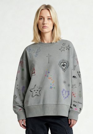 Blondine vrouw die een grijze oversized trui draagt met zwarte, kleurrijke krabbels in de stijl van sterren, vogels, hart, schedel, kruis en andere symbolen.