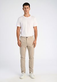 Pantaloni beige a vestibilità slim con un singolo bottone nero, abbinati a una t-shirt bianca a maniche corte e sneakers bianche.