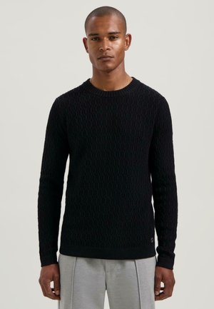 Schwarzer gestrickter Pullover mit strukturiertem Muster, Rundhalsausschnitt und langen Ärmeln. Gerippte Bündchen und Saum, kombiniert mit hellgrauen Hosen.