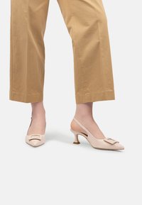 Pantofi cu toc kitten bej, cu detalii de cataramă pătrată, purtați cu pantaloni scurți bej pe un fundal alb.