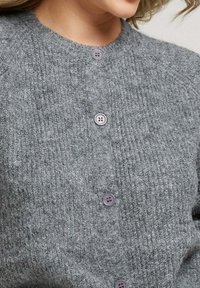 Gilet gris en tricot avec un col rond, un tissu texturé et quatre boutons ronds violets à l'avant pour la fermeture.