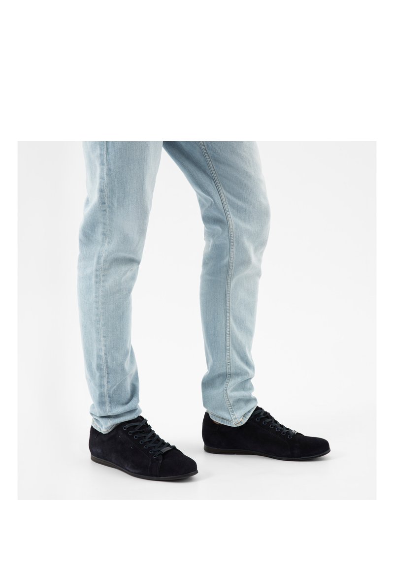 Persona che indossa jeans azzurri chiari e scarpe casual nere con lacci, in piedi con le gambe leggermente divaricate su uno sfondo bianco.