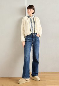 Chaqueta recortada de textura suave en color marfil sobre una camisa de botones a rayas. Combinada con pantalones vaqueros de pierna ancha y zapatos de plataforma en color beige.