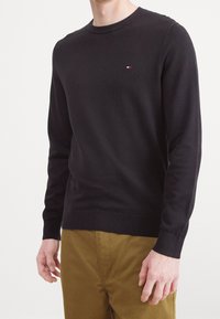 Pull noir en maille avec un col rond, des poignets côtelés et un petit logo sur la poitrine. Associé à un pantalon kaki, illustrant une tenue décontractée.