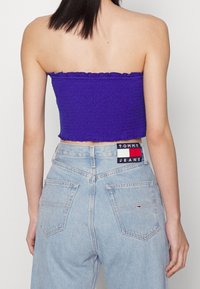 Haut cropped sans bretelles de couleur violette avec une texture smockée, associé à un jean en denim bleu clair taille haute avec un patch logo au niveau de la ceinture.