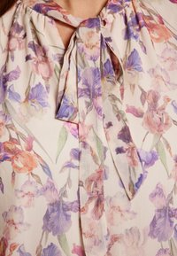Leichte, durchscheinende Bluse mit einem floralen Muster in Pastellfarben. Krawatten-Halsdetail und weicher Stoff mit einer fließenden, luftigen Textur.