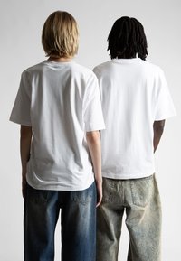 Dvaja jednotlivci nosia voľné biele bavlnené tričká. Jeden ich kombinuje s tmavomodrými džínami; druhý s bledými, svetlými džínami.