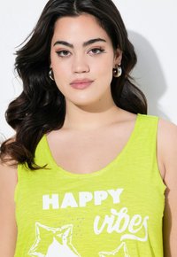 Top de tirantes verde lima con escote redondo, diseño sin mangas y texto blanco decorado con strass, exhibido sobre un fondo liso.