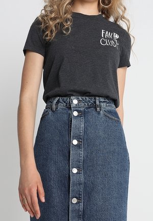 Woman wearing a dark gray "Fan Club" t-shirt tucked into a high-waisted, button-front denim skirt, with curly hair and hoop earrings.