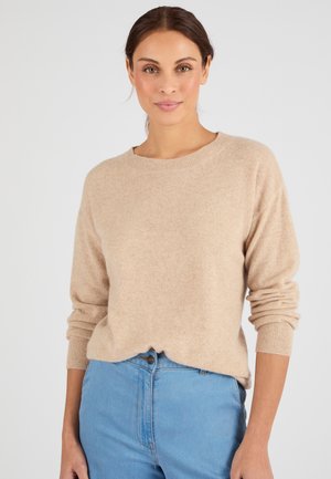 Frau mit braunen Haaren, die einen beigen Pullover mit langen Ärmeln und blaue hoch taillierte Jeans trägt, steht vor einem schlichten hellen Hintergrund.