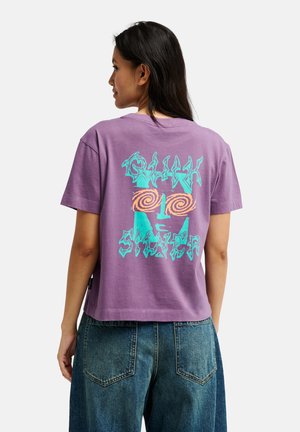 Vrouw met lang donker haar draagt een paarse T-shirt met een turquoise en oranje abstract gezicht en de tekst "Quik Silver" op de rug en blauwe spijkerbroek.