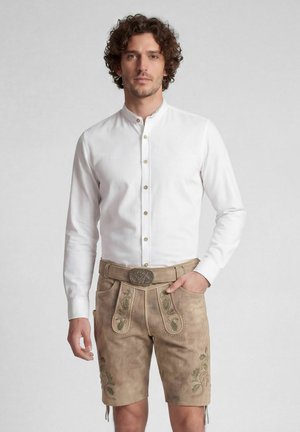 Mann mit lockigem Haar, der ein weißes Hemd mit Knöpfen und beige traditionelle Lederhosen mit grünen Stickereien trägt.