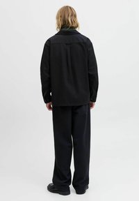 Schwarze Hemdjacke mit leicht oversized Passform, ausgestattet mit einem Kragen, Knopfleiste und einem Rückendetail mit Kellerfalte, kombiniert mit dunkler Hose.